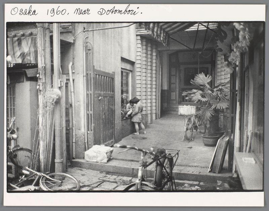 The image is a black and white photograph depicting an urban alleyway scene with the caption "Osaka 1960, near Dotombori." The setting appears to be in Japan during that era. In the foreground on the left side of the frame lies a discarded bicycle lying horizontally against what seems like a pillar or wall, its tires partially obscured by some debris and foliage.

In the middle ground, two young individuals are engaged with something outside a building's window; one is seated while the other stands beside them, leaning in. Their actions suggest they might be conversing or inspecting an object together. The alleyway leads to what seems like residential buildings on both sides, characterized by their windows and doors.

To the right of this central activity area are various items scattered along the ground: a folded parasol with its pole sticking up, potted plants including one resembling palm trees in large pots, and some stacked objects that could be household goods or tools. The scene is devoid of any vibrant colors due to the monochromatic filter.

The overall atmosphere evokes a sense of everyday life within an urban residential area during the late 1950s-early 60s in Japan, capturing both mundane details and human interaction as captured by Ed van der Elsken.