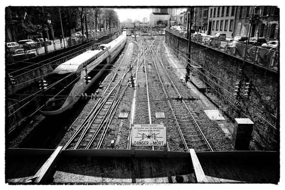 Black and white overhead view of a series of railroad tracks in between brick walls, with a train on the left-most track, and a small sign in the foreground reading "Danger de Mort"