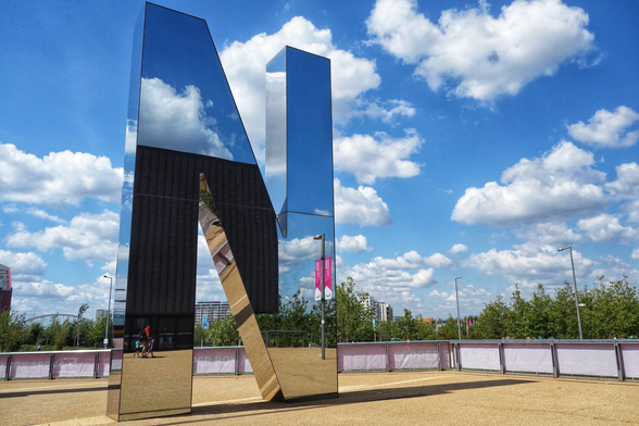 This image showcases a large, reflective metallic sculpture of the letter "N" from the "R U N" installation located in Queen Elizabeth Olympic Park, London. The sculpture stands prominently against a bright blue sky dotted with fluffy white clouds. The polished surface of the letter reflects the sky, clouds, part of the Copper Box Arena, a street lamp, people walking by, and surrounding greenery, creating a dynamic visual effect. The ground is covered in gravel, and the installation is enclosed by a low fence. In the background, trees and urban structures are visible, adding context to the park setting.