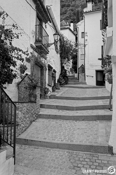 A narrow, winding street with stone steps and whitewashed buildings, adorned with potted plants. A vintage lamp hangs above, creating a charming, serene atmosphere in a picturesque setting.