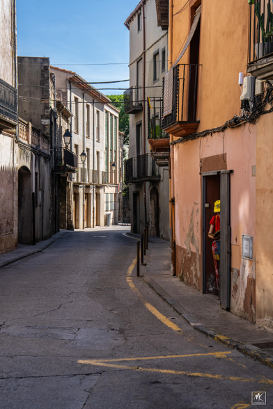 Color photo of a narrow curving street in a medieval Catalonian town with a partially obscured person with a yellow cap standing in a doorway. 
