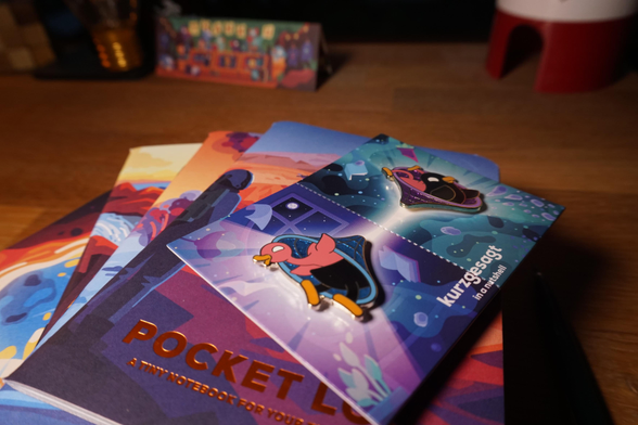 A picture of the wormhole pin, lying on top of 6 colourful notebooks. The notebooks are DIN A6 sized. They have space themed graphics on them. They are from the Mars and deep space collections by Kurzgesagt. 