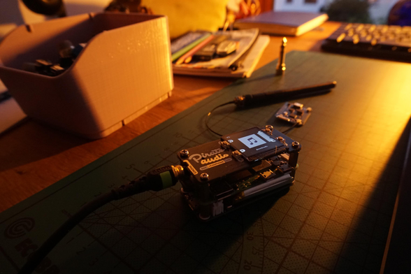 The main focus of the image is the Pi with the two HATs attached. The UPS HAT with its battery is connected from below using Pogo Pins. The Pirate Audio HAT has a display and 4 buttons and sits on top of the Pi. My headphones are currently connected to it. 
In the background there lay some notebooks, pens and other electronics.