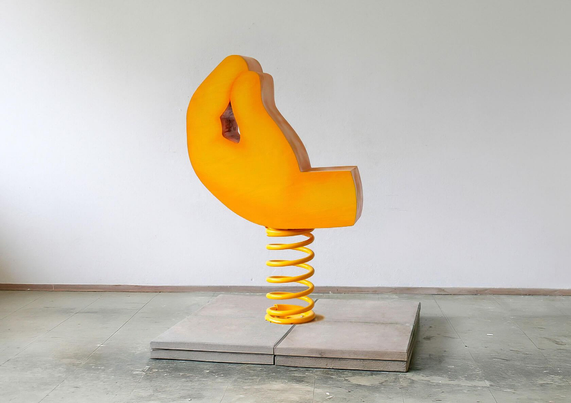 A bright yellow hand sculpture on a spring base, mounted on a gray platform, set against a plain wall.