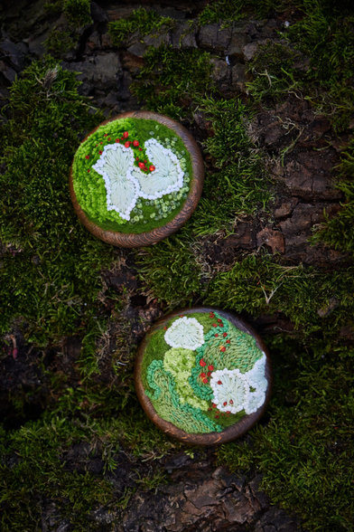 Two round and small hand-embroidered artworks rest on a moss-covered tree trunk. Each features green embroidery resembling moss, accented with softly glowing red dots. White, lichen-like shapes stand out against the green textures, while the wooden frames blend naturally with the earthy forest background.