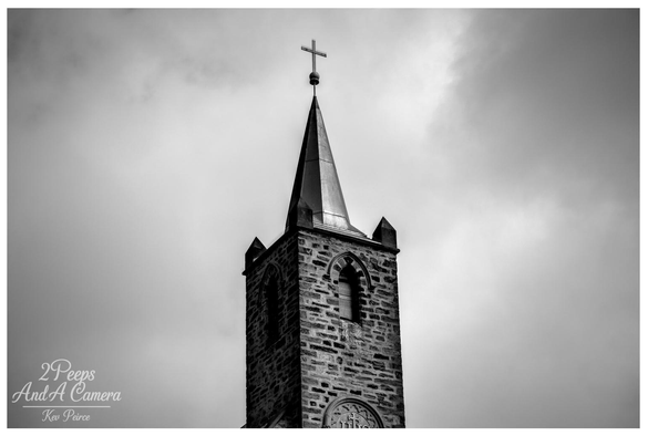 Black and white photograph showing the tall, stone steeple of a church in Burra, South Australia, topped with a cross. 

The spire is dramatic against a cloudy, atmospheric sky, emphasizing the architectural details of the bell tower.