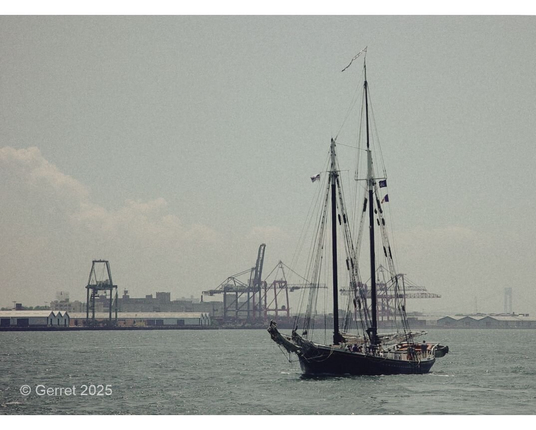 A vintage-style image showing a tall ship sailing on calm waters with industrial cranes onshore in the background. The setting feels nostalgic and serene.