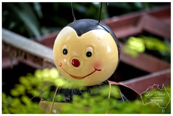 A close up, eye level photograph of a whimsical, spherical garden ornament shaped like a smiling ladybug or bee.  The ornament has a creamy yellow face, a black head, a small red nose, and black antennae. It is positioned low among lush green foliage, with blurred, rustic red and brown wooden structures visible in the soft focus background.