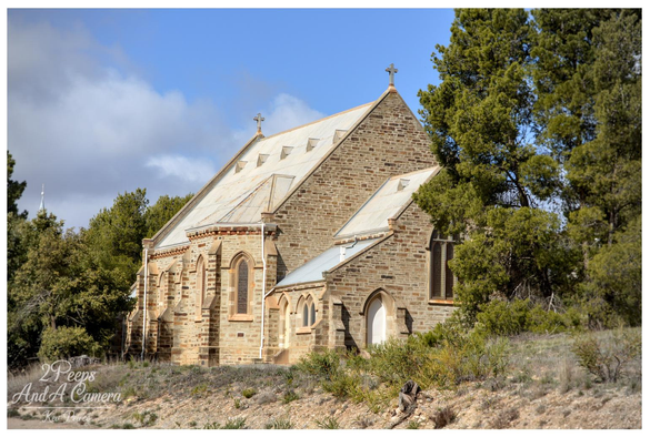 Photograph showing a historic stone church building on a slight incline under a bright blue sky with white clouds.

The church features Gothic style windows, a corrugated iron roof, and is flanked by tall, dense green pine like trees and dry scrub in the foreground.