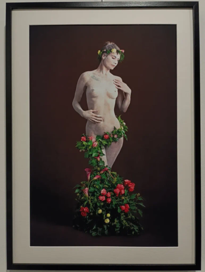 Photographie encadrée montrant une femme nue posant debout, le corps partiellement couvert de feuillage et de fleurs colorées, avec une couronne florale sur la tête, sur fond uni sombre.