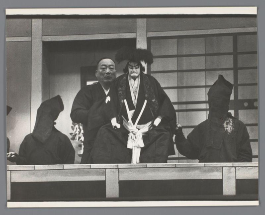 The image captures a group of individuals in traditional Japanese attire, possibly during a performance or cultural event. The central figures are two women dressed in elaborate kimonos with wide sashes and fur-trimmed hoods known as 'kakishibu'. They sit side by side on what appears to be an elevated platform against a backdrop resembling stage curtains. One woman has white makeup around her eyes, which is characteristic of certain traditional Japanese theater roles.

Surrounding these central figures are individuals wearing dark robes and head coverings that obscure their faces entirely. These garments suggest they could be performers or actors in the scene. The overall mood conveyed by the attire and setting suggests a formal cultural occasion with an air of solemnity. No discernible text is present, and the photograph has no explicit indication of its context beyond this detailed description.

This image provides insight into Japanese traditional theater practices, specifically those involving puppetry known as 'bunraku', which dates back to at least the late 16th century in Japan. The attire worn by both performers and audience members hints at their roles within the performance, reflecting historical customs where certain garments denoted specific characters or statuses.