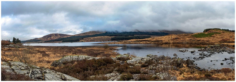 A panoramic view of a serene landscape featuring a calm lake surrounded by rolling hills and rocky terrain. The foreground includes rocks and sparse vegetation, while the background displays a cloudy sky over hills with patches of trees. The atmosphere is tranquil and natural.