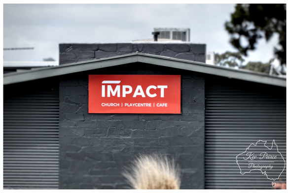 A bold red rectangular sign with the white text "IMPACT" is centered beneath the apex of a dark grey, gable-style roof.   Below "IMPACT" in smaller text are the words "CHURCH | PLAYCENTRE | CAFE". The building's walls feature both corrugated iron and dark, textured stone.