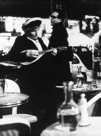  The image is a black and white photograph. In the center of the image, there is a person who appears to be a man playing an instrument. He is dressed in clothing that suggests a style from an earlier era, possibly mid-20th century, and he has his eyes closed, which might indicate he is performing or deeply engaged in the music. The setting is a bar or restaurant with tables and chairs visible in the background, suggesting a public dining area. There is also a bottle on one of the tables, further indicating that this is a social setting.

There are two other people present in the image; one appears to be standing near a table, possibly interacting with it, while the other person is sitting at a bar counter. The individual playing the instrument is slightly off-center and occupies a larger portion of the frame than the other people. The focus on this central figure suggests that he is the main subject of the photograph.

The image has a grainy texture which is characteristic of black and white photography, especially when it comes to capturing older scenes or vintage aesthetics. The overall composition of the image places the musician as the focal point against a background that includes other patrons in the establishment. 