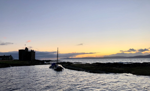 A tranquil scene featuring a castle silhouette against a sunset sky. A small boat is moored near a grassy shoreline, with water reflecting the soft hues of the evening twilight. In the background, distant hills and a calm sea complete the serene atmosphere.