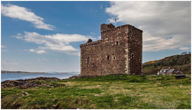 A historic stone castle stands near the water, surrounded by grassy land. A small house is visible in the background, with a partly cloudy sky overhead.