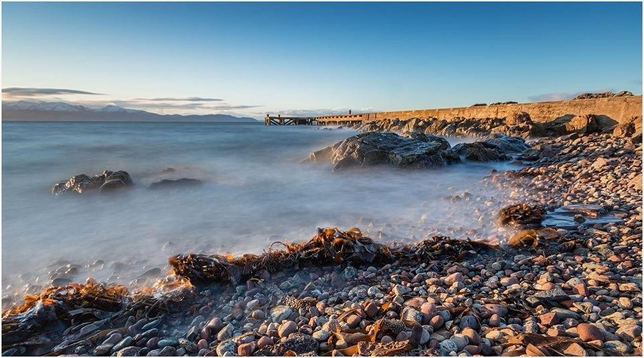 A serene coastal scene features a rocky shoreline with smooth pebbles and seaweed, leading to gentle waves. A wooden pier extends into the water, under a clear blue sky with distant mountains visible. The overall atmosphere is calm and picturesque.