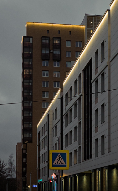 The photograph depicts an evening cityscape. In the center of the frame is a tall building with brown walls and large windows, illuminated from within. To its right is a shorter building with white walls and windows, also illuminated. Between the buildings is a pedestrian crossing sign depicting a person crossing the street. Other buildings and trees are visible in the background. It is late evening, the sky is gray.