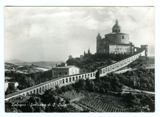  The image is a black and white photograph depicting a scenic landscape, primarily focusing on an old city with historical architecture. In the foreground, there's a prominent castle-like structure with multiple towers, resembling a fortress or bastion, perched atop a hill. The architectural style suggests it could be of European origin. Below this fortification, a long stone wall meanders through the landscape, creating a path that leads towards the viewer.

The image captures two distinct vantage points: one from a lower perspective looking up towards the fortress, and another from a higher vantage point providing a broader view of the city and its surroundings. The latter perspective shows a well-defined road or pathway leading downhill to what appears to be a town with modern buildings.

The image also includes textual elements: there are two lines of text at the bottom, each in a different language. The upper line is in Spanish, while the lower line is in Italian. The content of the text is not provided in this description, but it typically includes location information or historical context related to the scene depicted in the photograph.

The overall impression is that of a historically significant site, possibly a castle or fortification, situated within an old town, which might be of interest for visitors interested in history and architecture. The image evokes a sense of timelessness and grandeur, typical of historical landmarks. 