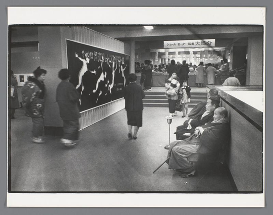  The image is a black and white photograph depicting an indoor scene that appears to be a museum or exhibition space. In the foreground, there are several people sitting on benches and standing around a central display. The individuals are dressed casually, and some are carrying handbags. They seem to be engaged in various activities, such as looking at exhibits or conversing with each other.

The central display features a large painting or mural that occupies a significant portion of the vertical space in the image. The painting appears to have a realistic style, but the details are not clear due to the distance and angle from which the photograph is taken.

Above the painting, there's an illuminated sign or information board with text that is too small and blurred to read clearly. To the right of the main photo, there is a smaller inset image showing the same scene but from a different perspective. This inset image captures more people who are not present in the main photo, suggesting it might have been taken at a different moment or angle.

The overall atmosphere of the image suggests an event or gathering taking place in a cultural or historical venue, possibly associated with art or education. The black and white color scheme gives the photograph a timeless or classic feel, emphasizing the contrast and textures within the scene. 
