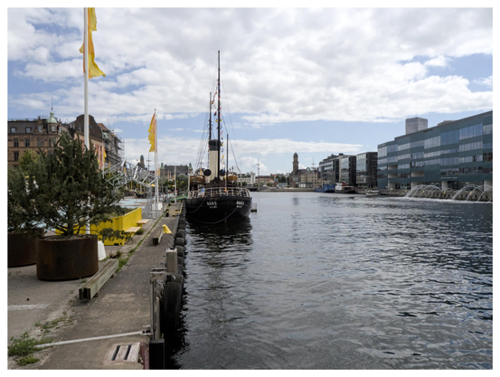 En liggande bild på ett svart och ljusgult fartyg som heter Bore. Hon ligger i en kanal. Två gula flaggor på vänstra sidan och fontäner med hus på högra sidan. Himmelen molnig med blå partier.