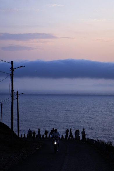Fotografía de una escena frente al océano. En la parte inferior una carretera gira hacia la izquierda por un acantilado, a la izquierda postes de luz se alejan en perspectiva. Asomandos en el arcen varias personas contemplan el panorama, una mujer se dirige hacia la cámara montando en bicicleta con la luz encendida, todo esto en tonos oscuros. En el centro del cuadro una masa de agua sobre la que hay el cielo con una nube que lo divide en horizontal, sobre la nube los colores mas calidos y claros bajo ella mas frios y oscuros.