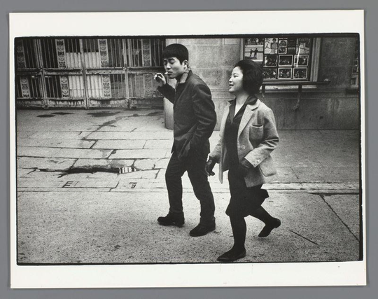 The image is a black-and-white photograph depicting two individuals walking side by side on a paved street. The person on the left is smoking a cigarette and is dressed in a dark jacket and pants, paired with dark shoes. The person on the right is wearing a light-colored coat over a dark outfit, with dark shoes, and is walking with a slight smile. They appear to be holding hands as they walk. The background includes a wall with a window, which has a grid-like pattern and what seems to be some posters or advertisements displayed on it. The ground is made up of large, uneven paving stones, and there is a visible crack or line on the pavement. The overall atmosphere of the image suggests a candid street scene, possibly from a past era given the style of clothing and the black-and-white format of the photograph.