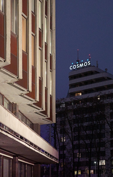 In the foreground is a section of an illuminated building with red panels. In the background, against the dark blue evening sky, a tall hotel is visible with the illuminated sign "Cosmos" on its roof.