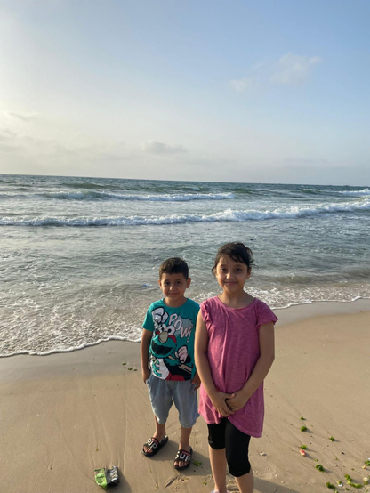 Two young children, a boy (Saji) and a girl (Aylan), stand close together on a sandy beach, the ocean waves visible behind them under a bright sky. The boy, wearing a teal t-shirt and shorts, smiles at the camera, while the girl in a pink shirt and black leggings looks forward with a smile, hands clasped.