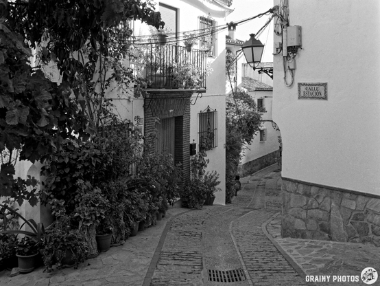 A narrow cobblestone street winding between whitewashed buildings adorned with plants and wrought iron detailing, leading to a sign that reads "Calle Estación,” reflecting a charming, quiet atmosphere.