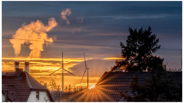 A sunset scene featuring puffs of smoke from industrial chimneys, wind turbines in the distance, and silhouetted rooftops and trees. The sky displays vibrant orange and blue hues.