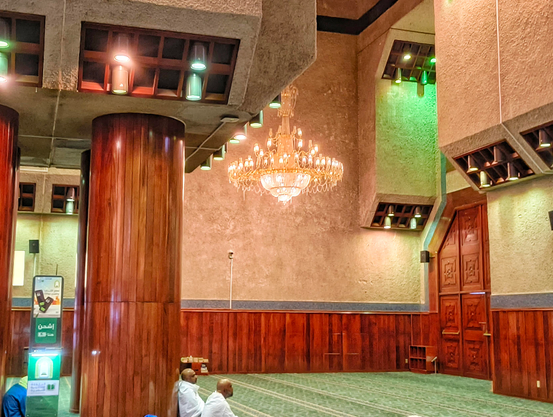 Interior view of a grand prayer hall in a Mosque featuring a striking blend of rough concrete ceiling structures and polished wood paneling on the lower walls and massive cylindrical columns. A large crystal chandelier hangs centrally, and geometric recessed lighting fixtures emit warm and green light. Two men wearing white Ihram pilgrimage clothing sit quietly on the green patterned carpet in the mid-ground. A large, intricately carved wooden door is visible on the right. A digital kiosk stands on the left.
