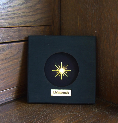 A small black shadowbox-style frame stands against a wooden corner. In the center is a circular window showing a glowing embroidered star made with fine gold thread. In its center shines a tiny light. A small plaque below the window reads ‘Lichtpuntje,' which means 'little point of light' or 'bright spot'.