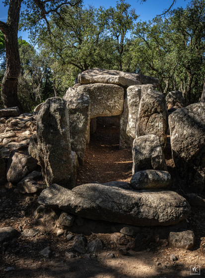 Color photo looking into the square opening of the central burial chamber of a prehistoric, megalithic stone dolmen structure with trees in the background. 