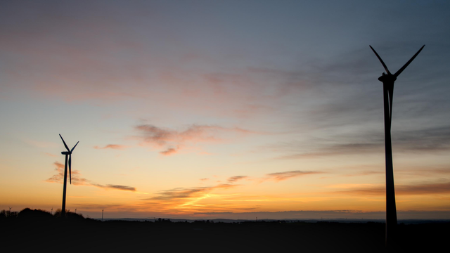 Sunset photo: two wind turbine silhouettes on a black ground frame the sunset gradient which goes from blue in the top to orange with dark clouds. In the upper third there is a pink cloud.