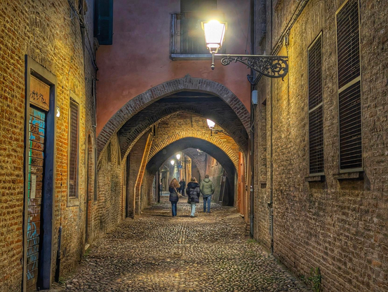A narrow cobblestone street in Ferrara framed by ancient brick arches and warm lamplight, with three people walking into the distance beneath the vaulted passageways that glow softly in the evening quiet.