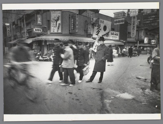 The image is a black-and-white photograph capturing a street scene, likely from the mid-20th century, judging by the clothing and architectural style. The setting appears to be an urban area, possibly in Japan, given the text on the signs and the architectural design.

In the foreground, a man in a long coat and hat stands prominently on the right side of the image. He is facing the camera and appears to be observing the street. Behind him, several people are walking, with some individuals carrying bags or baskets. The pedestrians are dressed in a variety of attire, suggesting a mix of casual and work-related clothing typical of the era.

The background is dominated by buildings with a mix of traditional and modern architectural elements. The buildings have numerous signs with Japanese characters, indicating businesses or shops. The signs are colorful and appear to be in bright colors, although the photo is in black and white. The street is paved, and there are some visible signs of wear, such as stains or discoloration on the pavement.

Overall, the image conveys a bustling street scene with a focus on the man in the foreground, who seems to be a central figure in the composition. The photograph captures a slice of daily life in a busy urban environment.