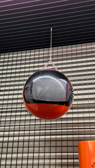 A spherical, red-orange TV set hanging on a chain in front of a metal lattice.