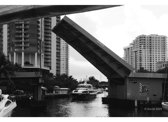 Black and white photo of a raised drawbridge over a waterway. A yacht passes beneath, amidst tall city buildings, conveying a bustling urban atmosphere.
