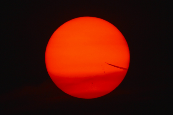 Photograph of a large red sun against a black background with a jet and contrail crossing into it from the lower right.