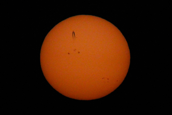 Photograph of a large yellow sun against a black background with a jet and contrail in the upper left crossing straight upwards.