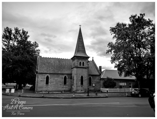Black and white photograph of the historic Ross Uniting Church in Tasmania, featuring its central stone tower and spire. 

The sandstone building is set at a street corner, framed by large, dark trees on the left and right, with a white picket fence visible on the right. The sky is heavily clouded.