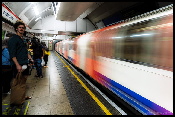 Photograph of passengers waiting on Oxford Circus Underground station platform with a red and white London Underground train with blue stripe blurred in motion passing through the station, fluorescent ceiling lights, yellow and black tactile warning strip and yellow safety markings visible on platform edge, metal mesh panels and white station infrastructure visible.
