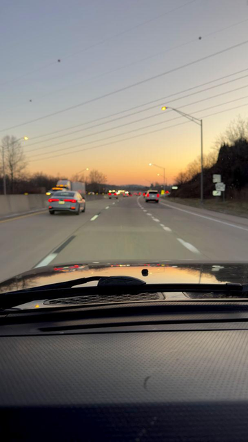 View through the windshield of a Toyota FJ Cruiser driving south on a multi-lane highway at dusk near the Lancaster-Berks county line. The sky glows orange along the horizon, fading into pale blue above. Headlights and taillights stretch into the distance, streetlights are just turning on, and the vehicle’s hood reflects the sunset's colors.