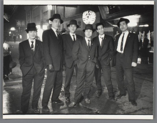 The image is a black and white photograph featuring six men standing together on what appears to be an urban street at night. They are all dressed in suits, ties, and hats that suggest they might belong to the same organization or group, possibly of professional nature due to their formal attire. The background shows blurred lights and signs indicative of nightlife, with one sign displaying Asian characters suggesting this scene is set in a Japanese city. There's no clear indication of any specific event taking place; it seems like a candid shot capturing these individuals together during an evening outing or social gathering.

Additional information about the image can be found on the provided source link: "Groepsportret van onbekende yakuza-mannen, Osaka" which translates to "Group portrait of unknown Yakuza men, Osaka." This suggests that the photograph may have been taken in Japan and features individuals who are likely associated with organized crime groups known as 'Yakuza.' The photographer is Ed van der Elsken, an acclaimed Dutch documentary photographer. His works often capture street life and culture, particularly focusing on East Asia during his time there.
