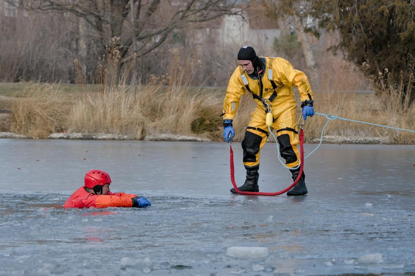 A color landscape photo of two men practicing an ice rescue. A man in an orange dry-suit is waiting calmly floating shoulder high in a hole in the ice of a lake. He has his arms extended onto the unbroken ice to hold his head above water. A man in a yellow dry-suit, with blue gloves, and black boots cautiously approaches the man in the ice water from the right. The second man is pulling along a rope with a clasp. There are brown bushes on the shoreline in the background.