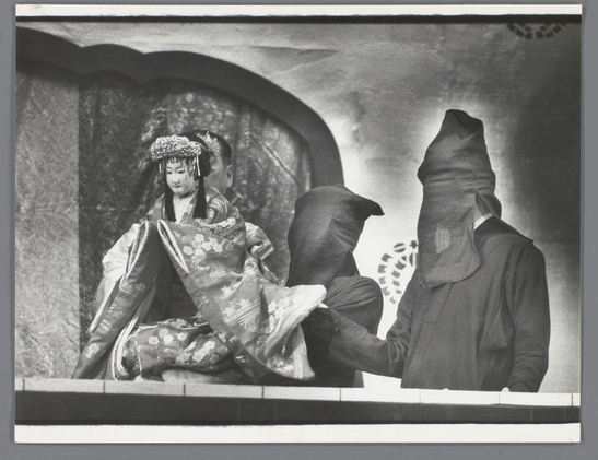 The image is a black and white photograph featuring three individuals on stage, likely part of a Bunraku puppet theatre performance. On the left side sits an individual dressed in traditional Japanese attire with ornate headpieces, possibly portraying a female character from historical or literary narratives common to such performances. This figure's posture suggests they are either engaging in conversation or presenting something.

On the right, two figures stand facing away from the camera and each other; both have their faces concealed by dark puppets' heads, typically used for puppetry within Bunraku theatre. The standing individuals wear what appears to be Western-style suits with visible patches of fabric suggesting a worn-out texture. Their stance is somewhat hunched, which may indicate contemplation or anticipation.

The setting seems simplistic, perhaps an interior stage setup indicated by the partial view of a draped wall in the background and a hint of architectural detailing above it. The lighting casts shadows that add to the dramatic effect often sought after in theatre performances like Bunraku.

This photograph captures not just the performers but also offers insight into their roles within this theatrical tradition, showcasing both costume details as well as puppetry elements used in live performance art from Japan during a time when such traditions were actively documented.