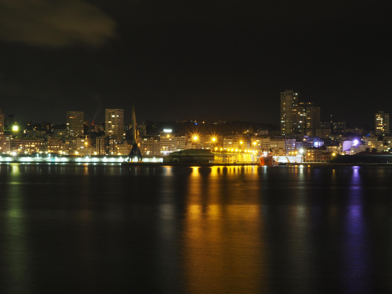 Foto noctura do Porto da Coruña, no centro da imaxe podemos ver as luces douradas do Peirao de Calvo Sotelo, a esquerda as luces brancas do Peirao do Centenario e por enriba deles no fondo vemos o "skyline" da cidade. Mais cerca de nos vemos as augas oscuras do porto iluminadas con reflexos.