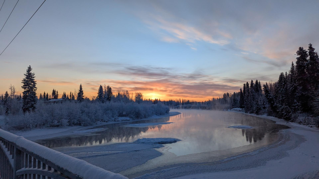A picture of a sunset across a river in the process of freezing. The trees on the left are completely covered in frost and snow while the trees on the right have yet to be covered