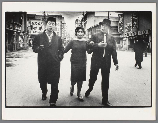 The image is a black and white photograph showing three individuals walking down an urban street. On the left, there's a man wearing a dark coat with a light-colored scarf around his neck; he appears to be leading the way as he holds onto the arm of another person in the center. The central figure seems to be guiding or supporting the third individual on the right, who is dressed formally and carries an item under their arm that could possibly be a cigarette case. Behind them are various storefronts with signs written in Japanese script, indicating this scene likely takes place in Japan during the time period suggested by the style of clothing and the overall atmosphere.

The individuals have distinctive attire: the man on the left wears casual streetwear from an earlier era, while the central woman sports a dress that suggests mid-20th-century fashion. The person to her right is dressed more formally with a suit coat and hat. Their expressions are neutral as they stride down what appears to be a busy commercial area.

Additional context for this photograph can be found in its accompanying information: it's part of the Ed van der Elsken collection, dated between 1959-60, depicting Kamagasaki, Osaka. This indicates that Jonge vrouw loopt tussen twee jonge mannen is more than just a casual snapshot; it captures a specific time and place with cultural significance in Japan during an era when Japanese society was undergoing significant change.